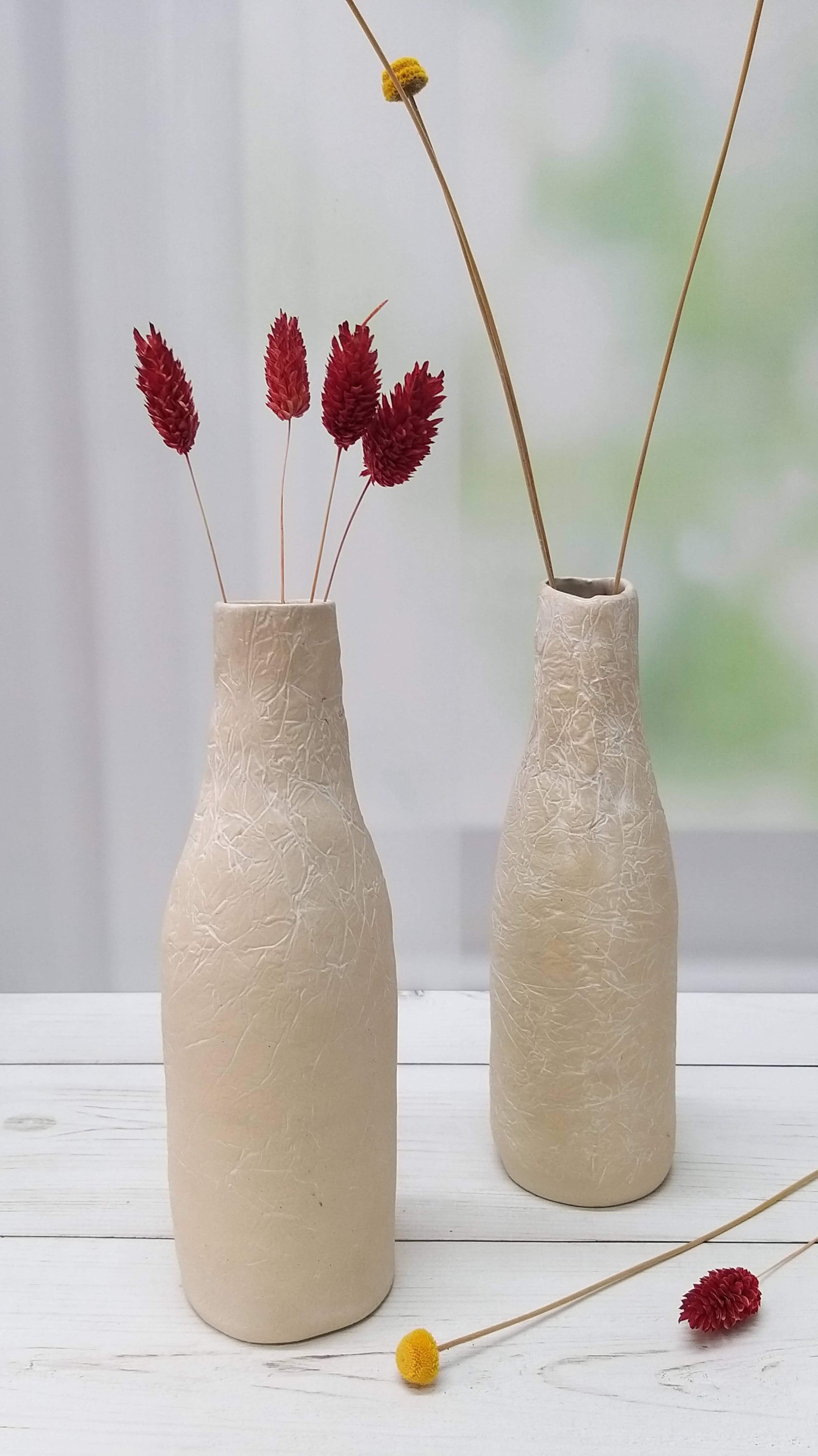שני אגרטלי קרמיקה לבנים בצורת בקבוק מונחים זה ליד זה ובתוכם פרחים אדומים וצהובים